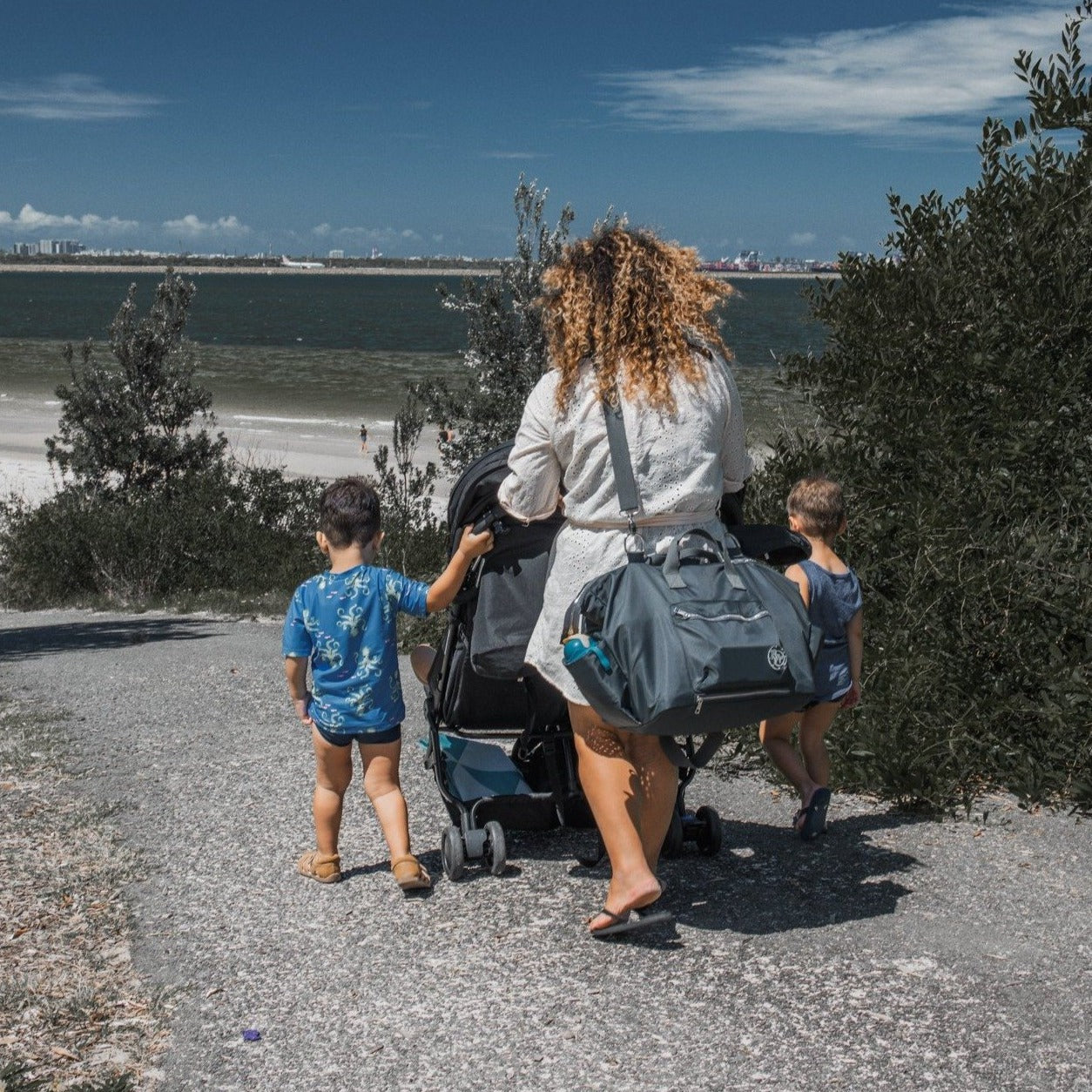 alt="mum pushing a pram wearing The DUFFLE Nappy Bag crossbody with toddlers at beach"