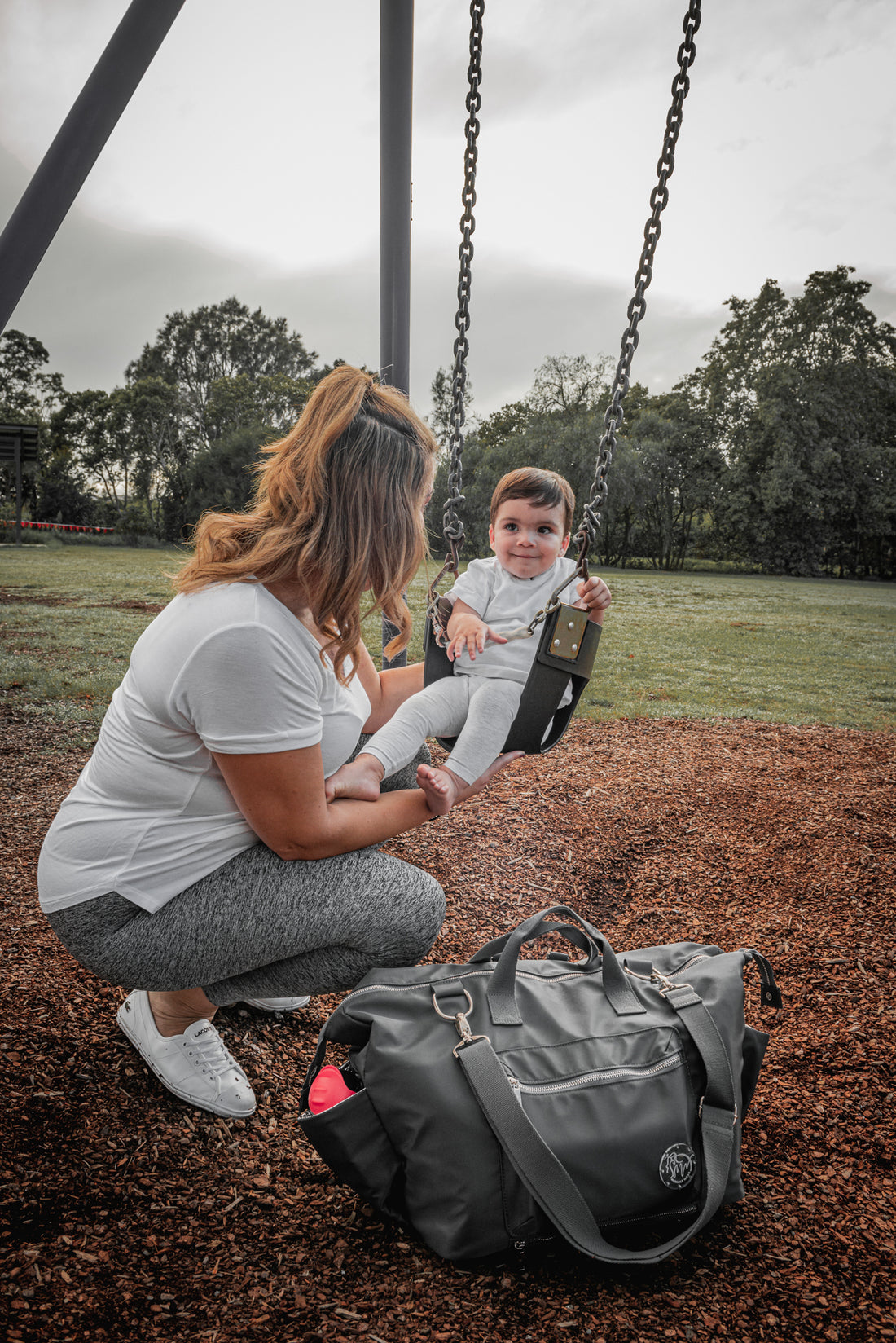 Mum kneeling in front of her baby boy who is sitting on the swing at a park playground. Beside them is The DUFFLE Nappy Bag in Chaos + Calm (charcoal).