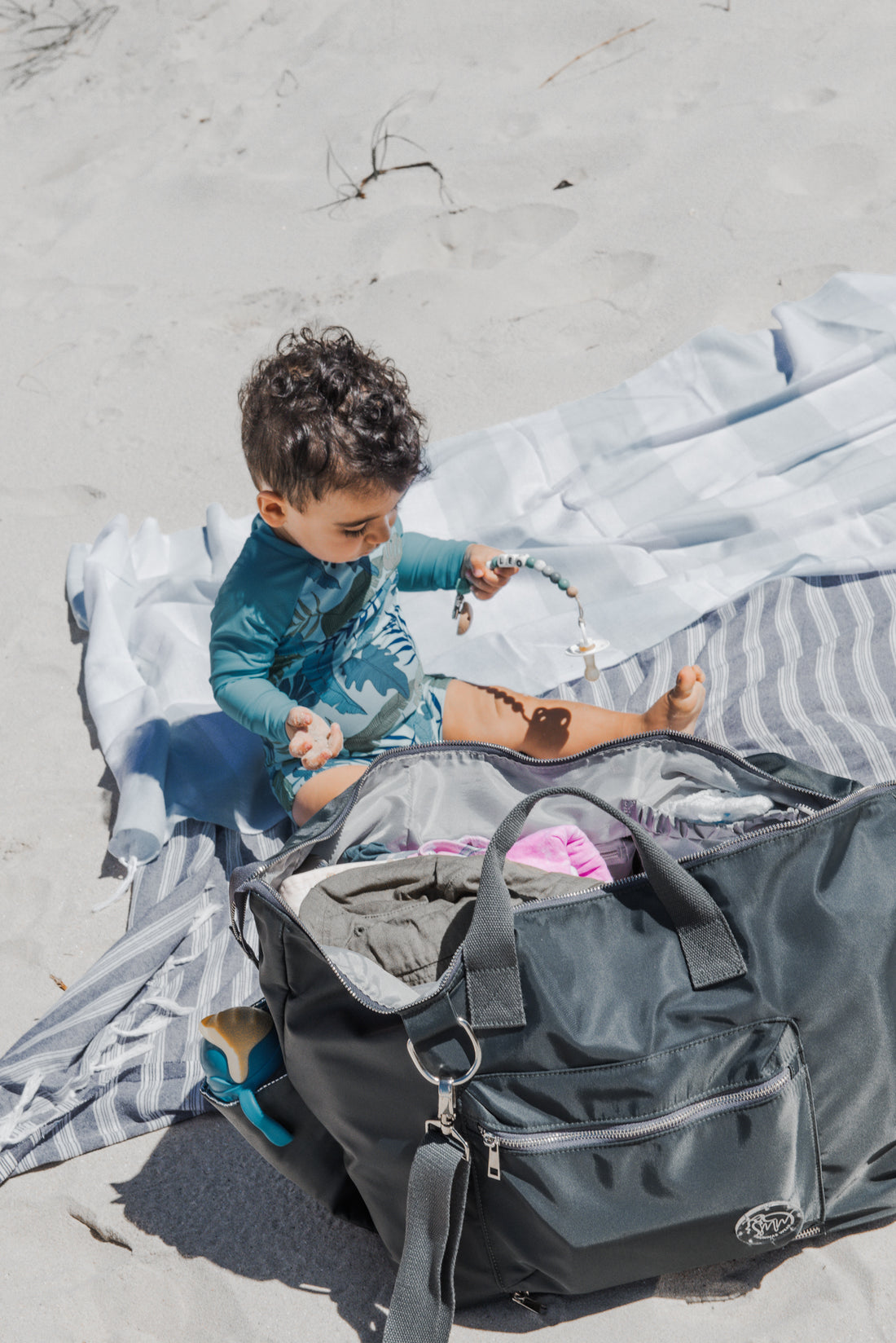 The DUFFLE Nappy Bag in Chaos + Calm (charcoal) sitting on beach towel next to baby boy.